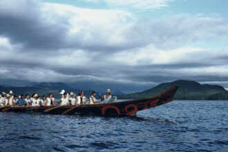 Haida hereditary chief Gidansda (Guujaaw) leads a group of paddlers aboard Luu Taas (“Wave Eater”), a 15-meter red-cedar canoe designed by acclaimed Haida artist Bill Reid for the 1986 World Exposition on Transportation and Communication held in Vancouver, British Columbia. Carving and paddling cedar canoes is one example of how Haida people are inextricably linked to both land and sea. Credit: Courtesy of Guujaaw